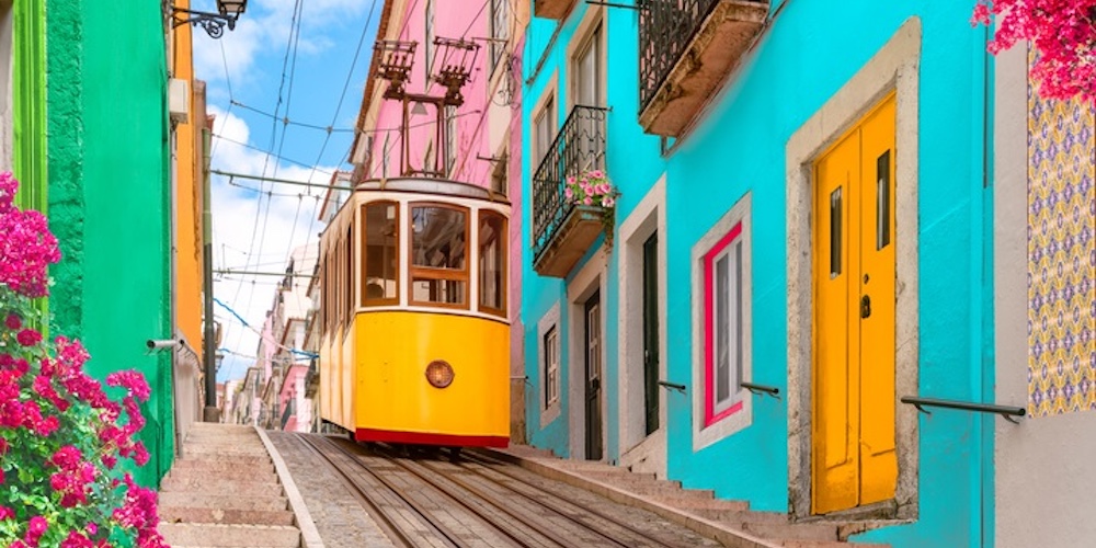 Yellow electric tram on a street with colorful houses and flowers on the balconies - Bica Elevator going down the hill of Chiado in Lisbon, Portugal.
