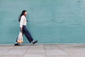 A woman walking with large strides infront of a large blue brick wall carrying a bag of shopping