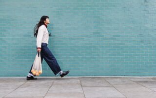 A woman walking with large strides infront of a large blue brick wall carrying a bag of shopping