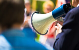 Man holding a megaphone on picket line while speaking to a crowd.