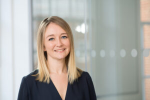 Portrait of a smiling blonde woman wearing a navy blazer in a bright office setting.