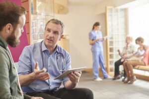 male doctor speaking to male patient. in the back ground a female helath worker speaking to 2 women in a waiting area