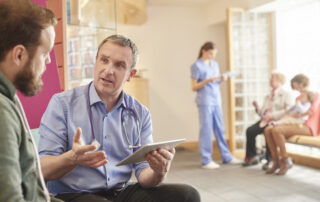male doctor speaking to male patient. in the back ground a female helath worker speaking to 2 women in a waiting area