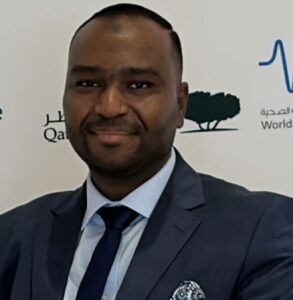Dr Hassan Farhat - BMJ Group - BMJ Group Portrait of a man in a dark suit and blue tie posing at a backdrop with logos, suggesting a formal event or conference.
