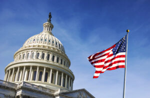 U.S. Capitol dome with an American flag waving on a clear blue sky.