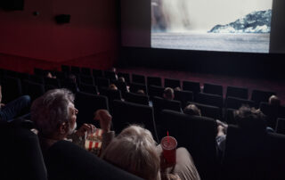 Audience seated in a dark cinema watching a snowy landscape on the large screen; foreground shows popcorn and drinks.