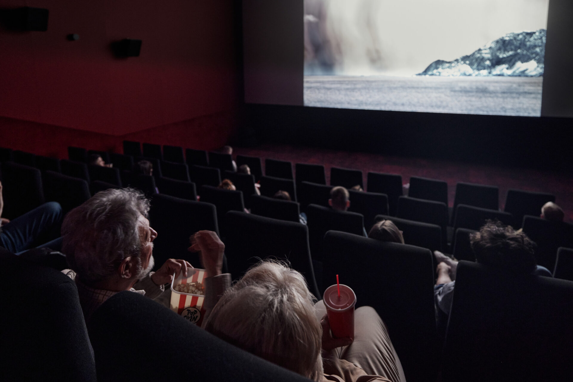 Audience seated in a dark cinema watching a snowy landscape on the large screen; foreground shows popcorn and drinks.