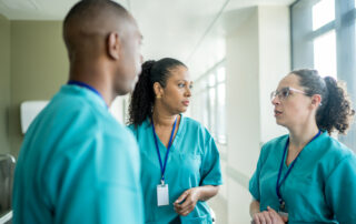 Three healthcare professionals in teal scrubs standing in a bright hospital corridor, deep in discussion.