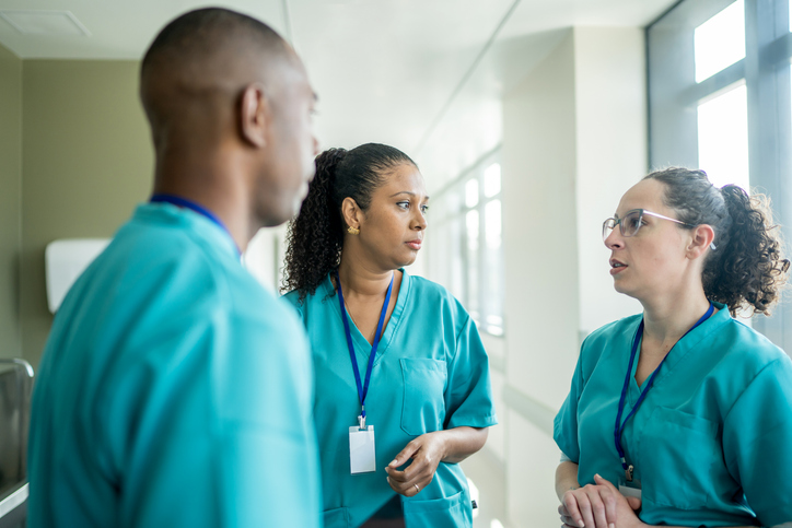 Three healthcare professionals in teal scrubs standing in a bright hospital corridor, deep in discussion.