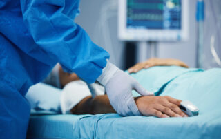 Nurse in blue scrubs and gloves checks a patient’s hand with a pulse oximeter while a monitor shows vital signs in the background.
