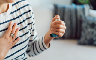 Person in a striped shirt uses a blue inhaler for asthma, pressing the device with one hand while touching their chest with the other.