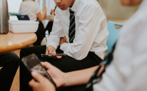 Young person in a white shirt and striped tie sits at a table, focused on a smartphone in a cafe-like setting with others in the background.