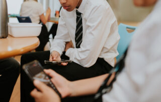 Young person in a white shirt and striped tie sits at a table, focused on a smartphone in a cafe-like setting with others in the background.