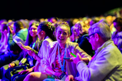 Smiling woman with a lanyard at a colorful night event, surrounded by cheering audience under neon lighting.