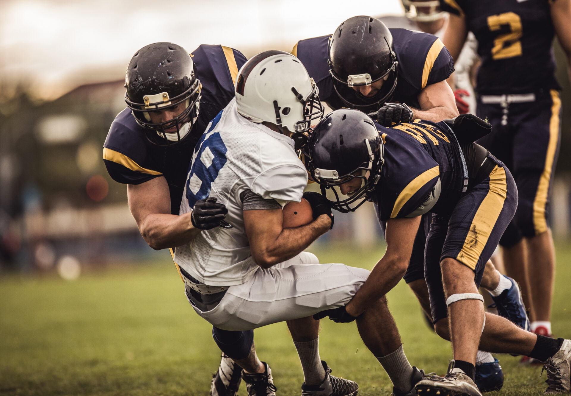 Rugby players in navy and gold uniforms collide in a tightly packed scrum on a grassy field, white jersey visible in the center.