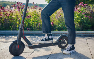 Person wearing black Converse sneakers and rolled jeans stands with one foot on a black electric scooter outdoors, with flowers in the background.