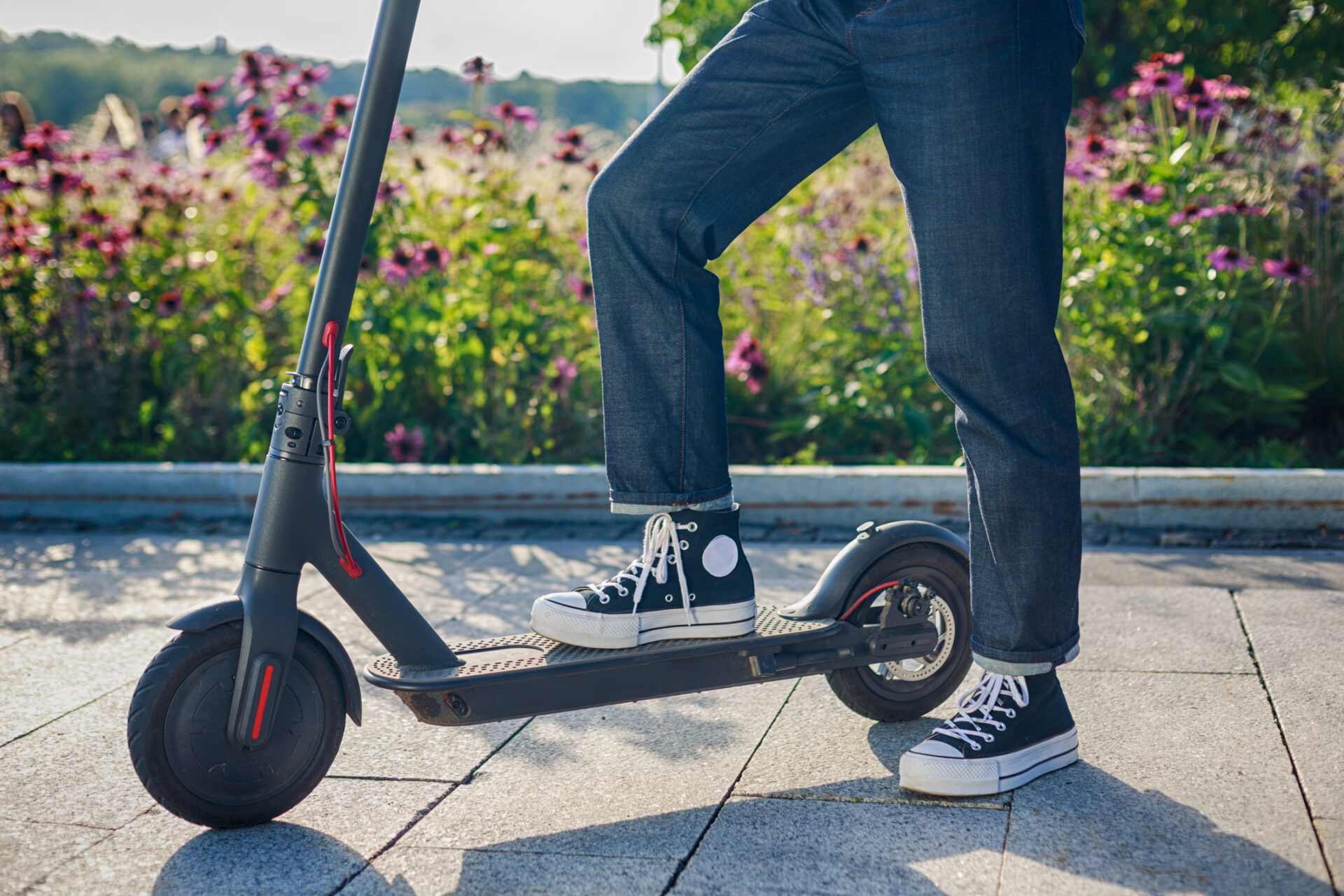 Person wearing black Converse sneakers and rolled jeans stands with one foot on a black electric scooter outdoors, with flowers in the background.