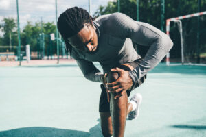 Athlete in a gray textured shirt bending to stretch his leg on an outdoor sports court.