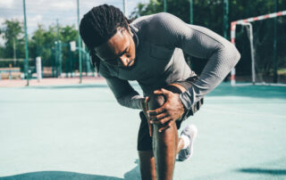 Athlete in a gray textured shirt bending to stretch his leg on an outdoor sports court.