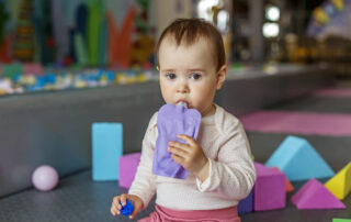 Toddler girl sitting among colorful foam blocks, sipping from a purple pouch.