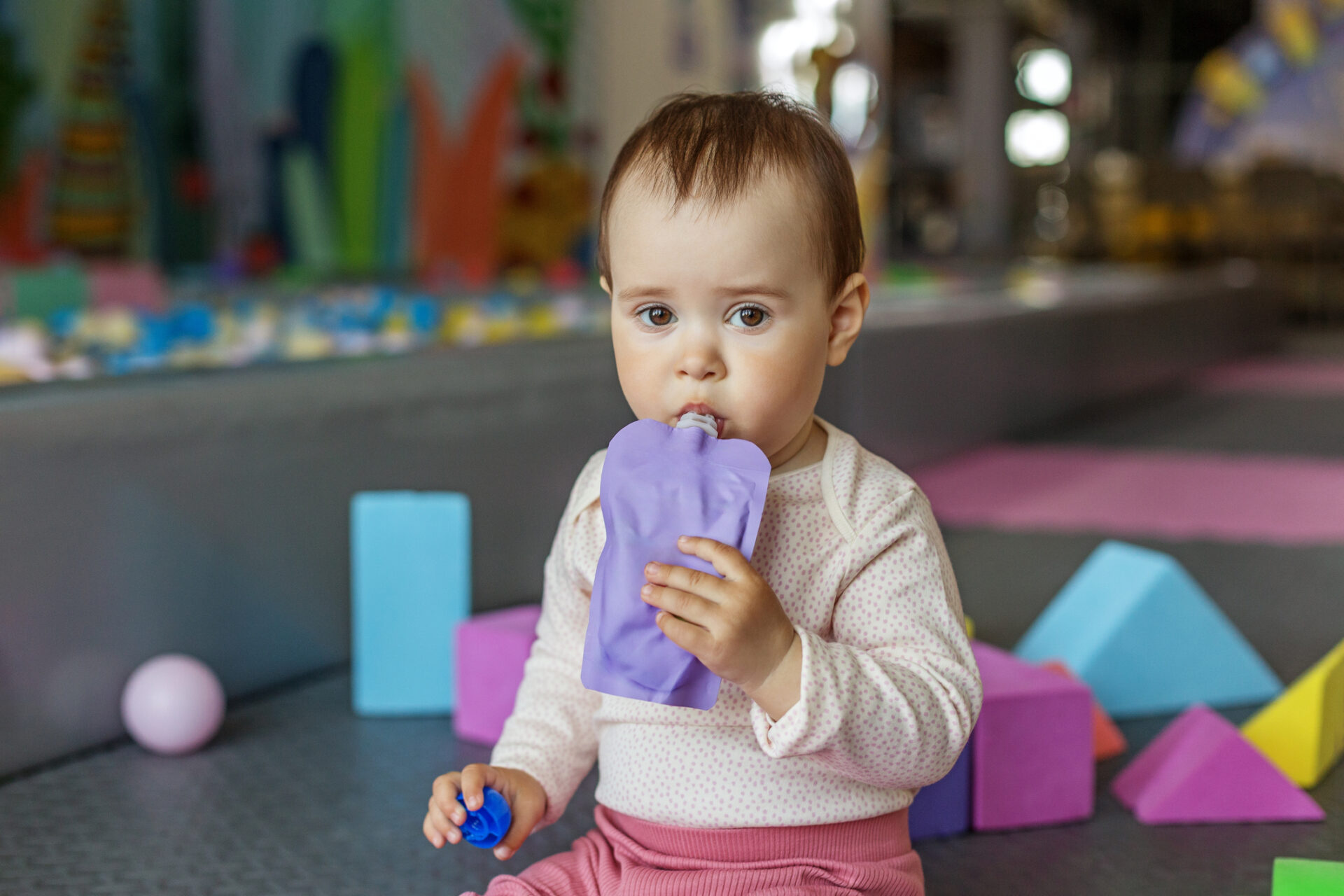Toddler girl sitting among colorful foam blocks, sipping from a purple pouch.