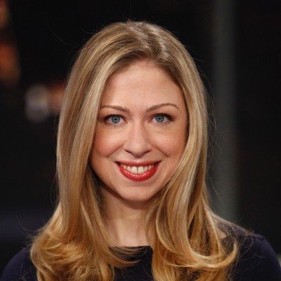 Head-and-shoulders portrait of a smiling blonde woman with blue eyes, wearing a dark top against a dark blurred background.