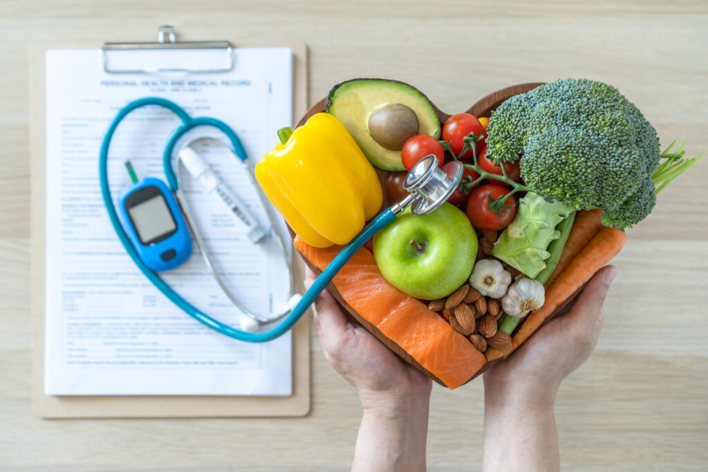 Hands hold a bowl of fresh vegetables and fruit (broccoli, avocado, peppers, tomatoes, apple, carrots) with a stethoscope and clipboard in the background.
