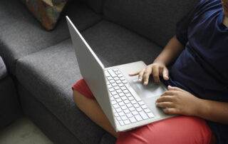 Child sitting on a gray couch using a silver laptop on their lap.
