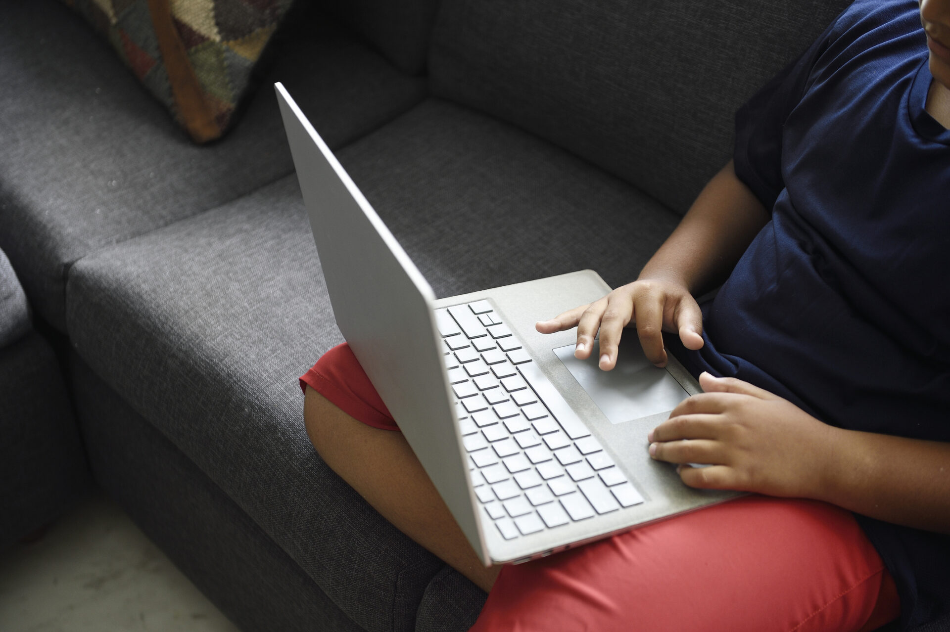 Child sitting on a gray couch using a silver laptop on their lap.