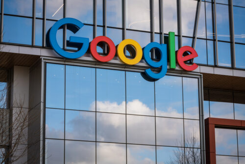 Google logo displayed on the glass facade of a modern office building with blue-sky reflections.
