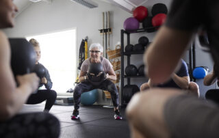 Older woman in a pink top squats while holding a black medicine ball during a group fitness class in a gym.