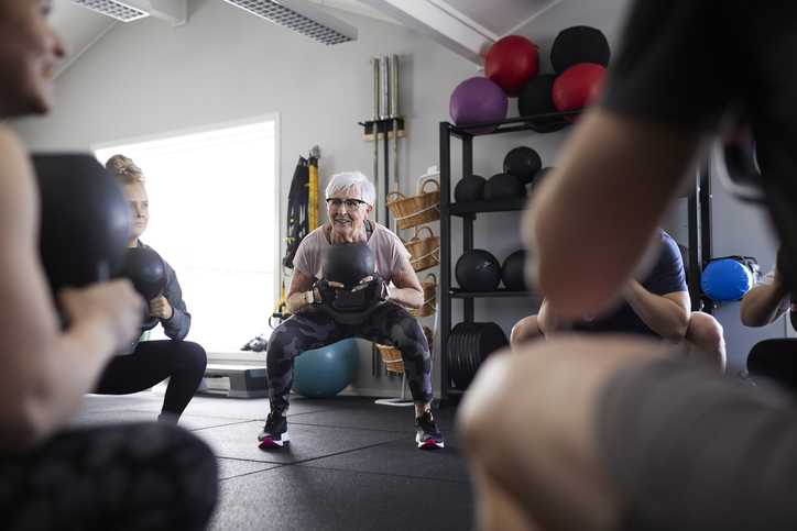 Older woman in a pink top squats while holding a black medicine ball during a group fitness class in a gym.