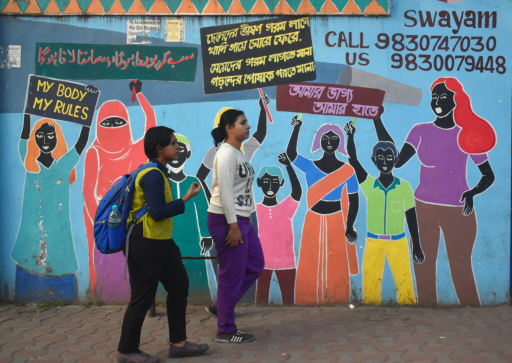 Two women walk past a vibrant mural of protest silhouettes holding signs, including one reading 'MY BODY MY RULES'.