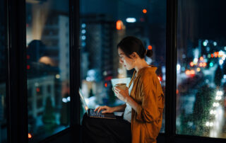 Woman typing on a laptop by a window at night, holding a mug; blurred city lights outside the glass.