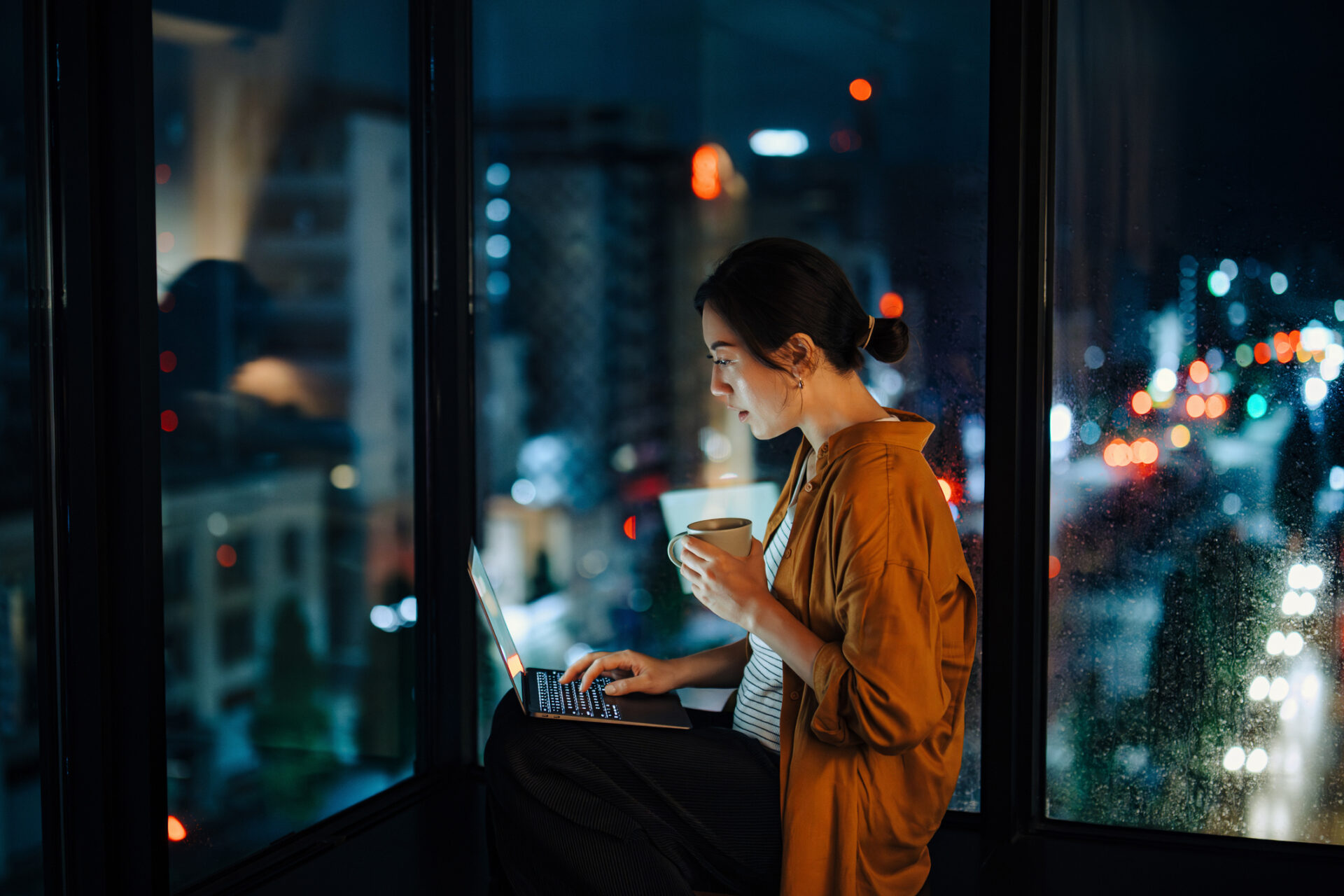 Woman typing on a laptop by a window at night, holding a mug; blurred city lights outside the glass.