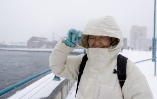 Person in a light-colored winter coat with hood up and turquoise gloves shielding eyes against cold wind on a snowy waterfront bridge, cityscape in background