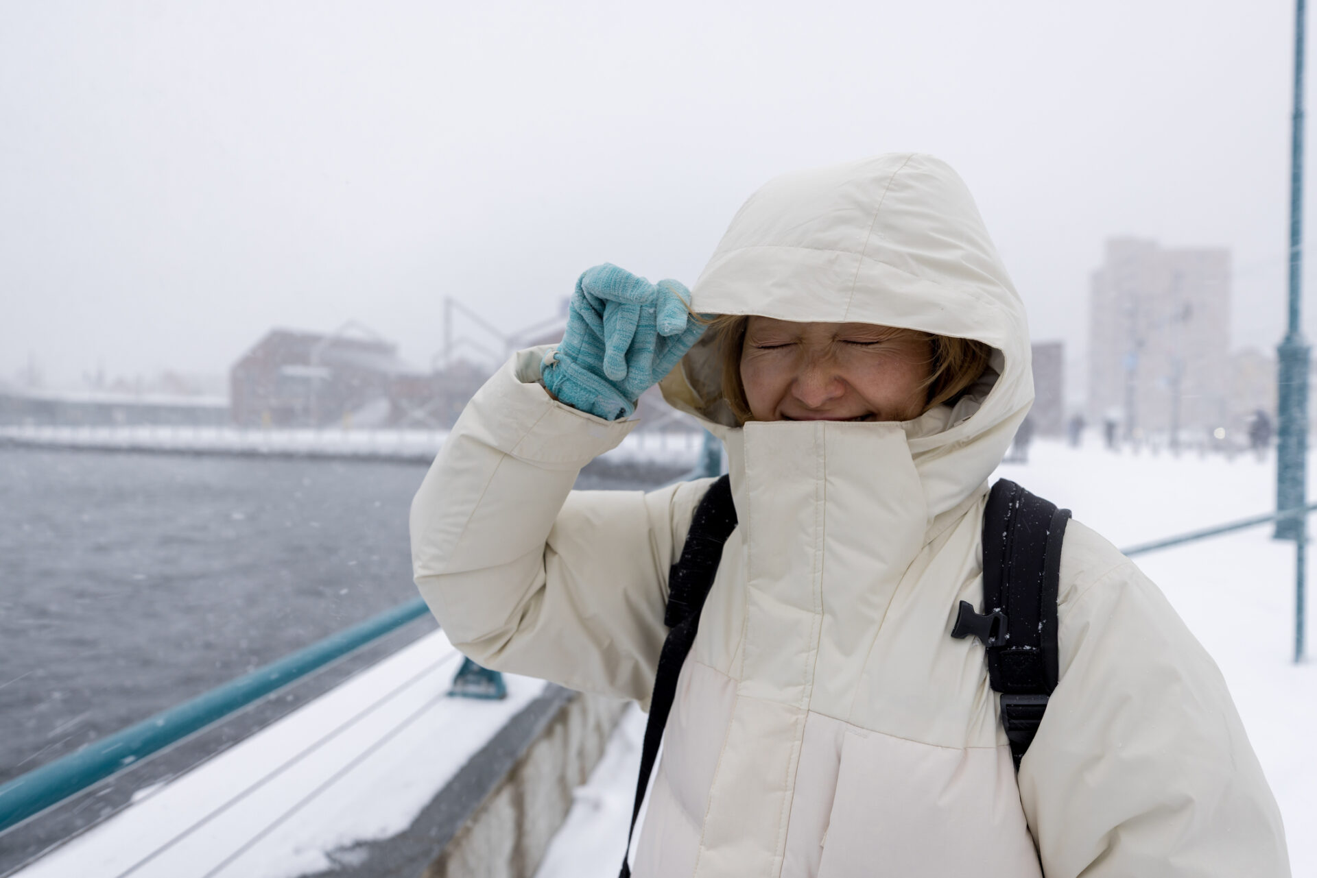 Person in a light-colored winter coat with hood up and turquoise gloves shielding eyes against cold wind on a snowy waterfront bridge, cityscape in background