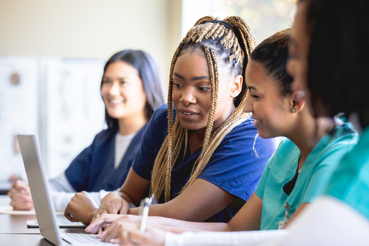 Three students sit at a desk in a classroom, collaborating on laptops during a study session.