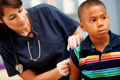 Nurse administers a vaccine to a young boy's upper arm; stethoscope around her neck in a clinic setting.