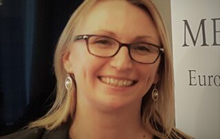 Smiling blonde woman with rectangular glasses, wearing a dark blazer and earrings, at a conference or event.