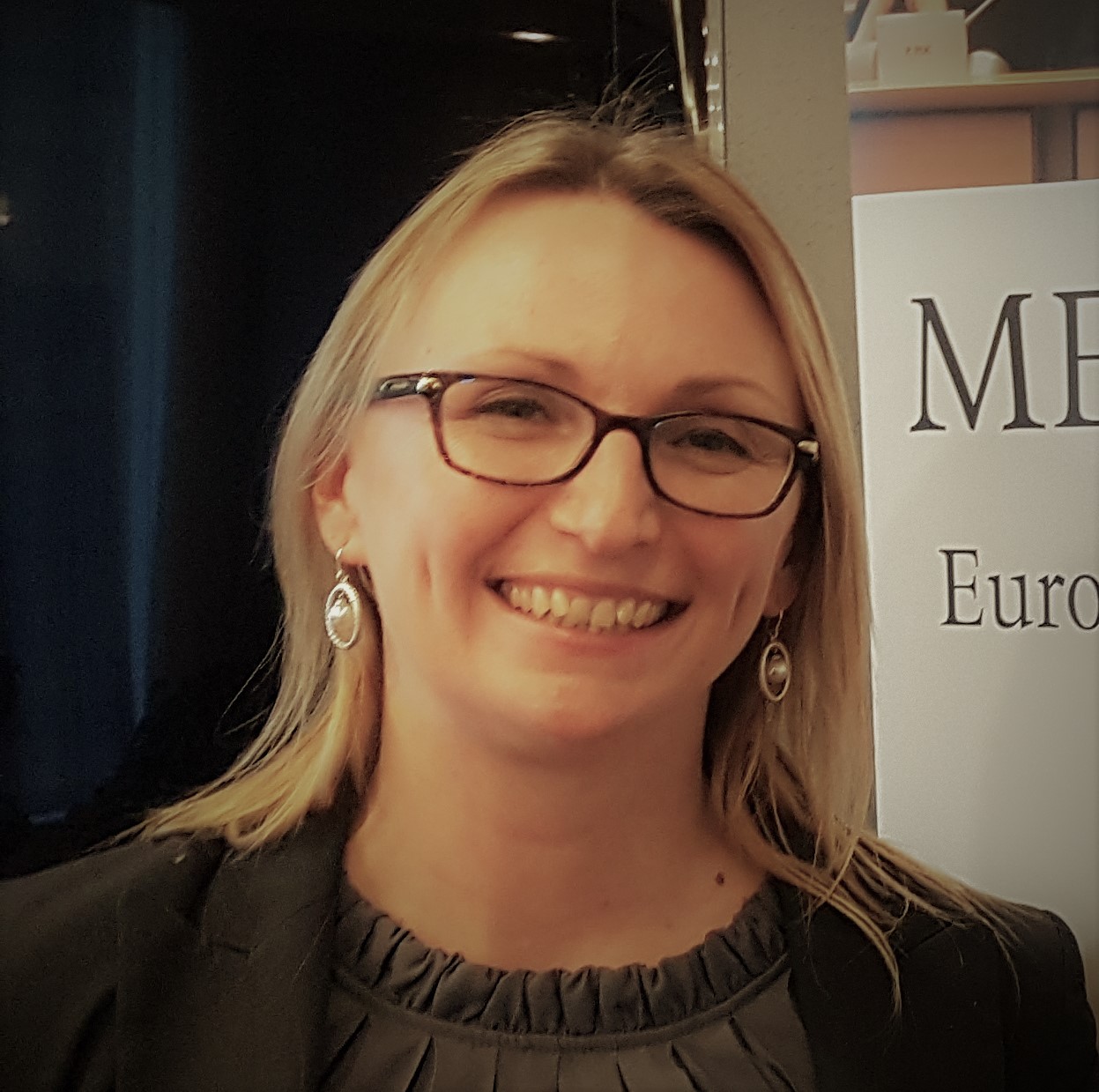Smiling blonde woman with rectangular glasses, wearing a dark blazer and earrings, at a conference or event.
