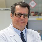 Male scientist wearing a white lab coat and glasses, looking at the camera in a laboratory setting.