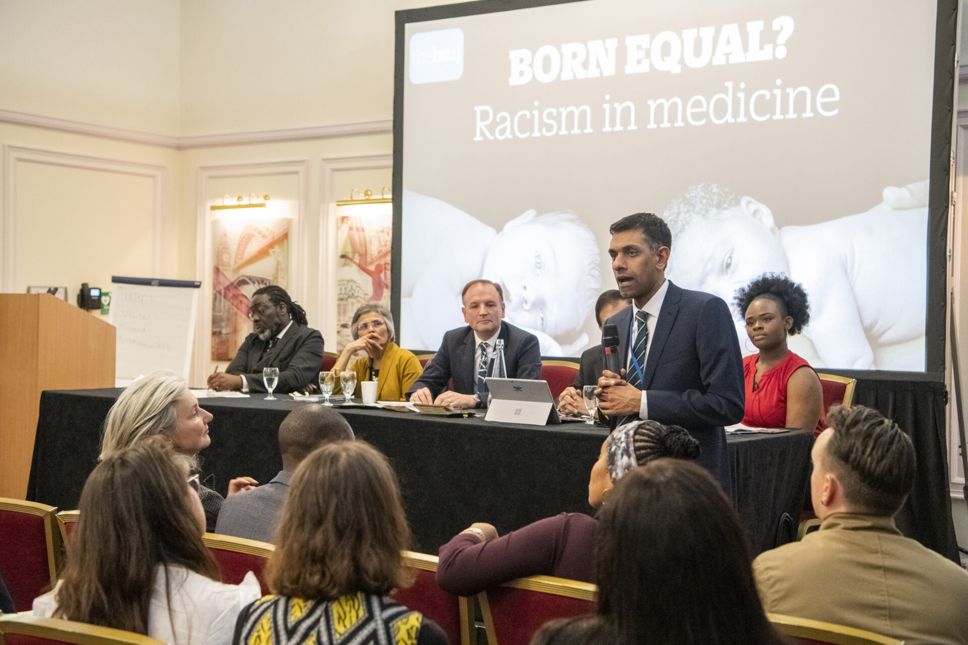 Panel of five seated at a long table on a stage, one man in a suit speaking into a microphone; large screen behind reads 'BORN EQUAL? Racism in medicine'