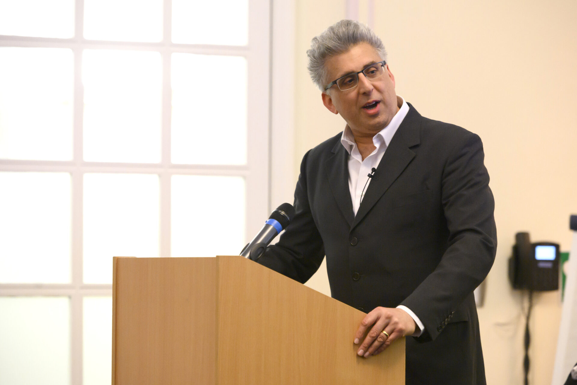 Man in a dark suit speaks at a wooden podium with a microphone in a bright room
