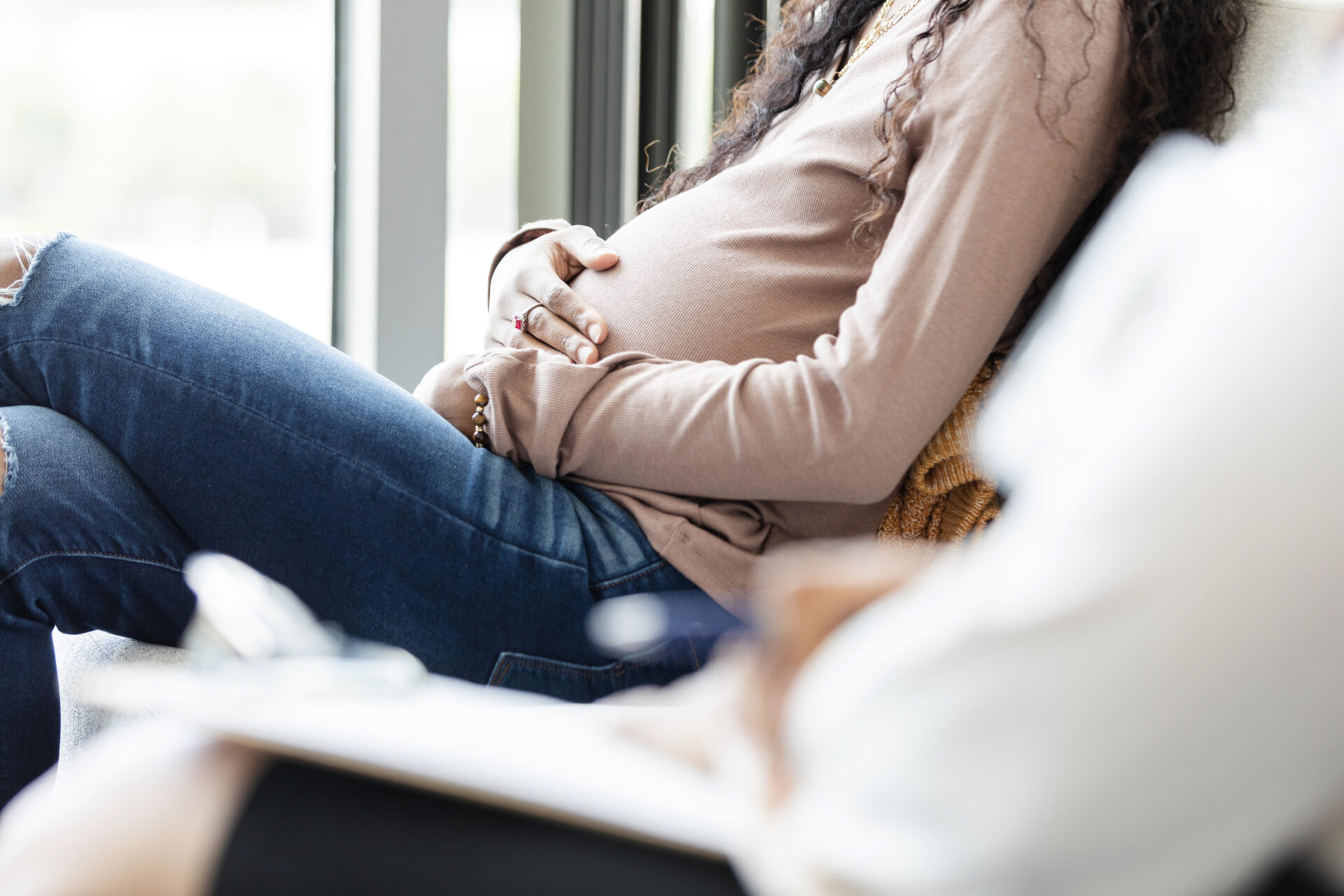 Pregnant woman sits by a window, hands resting on her rounded belly.
