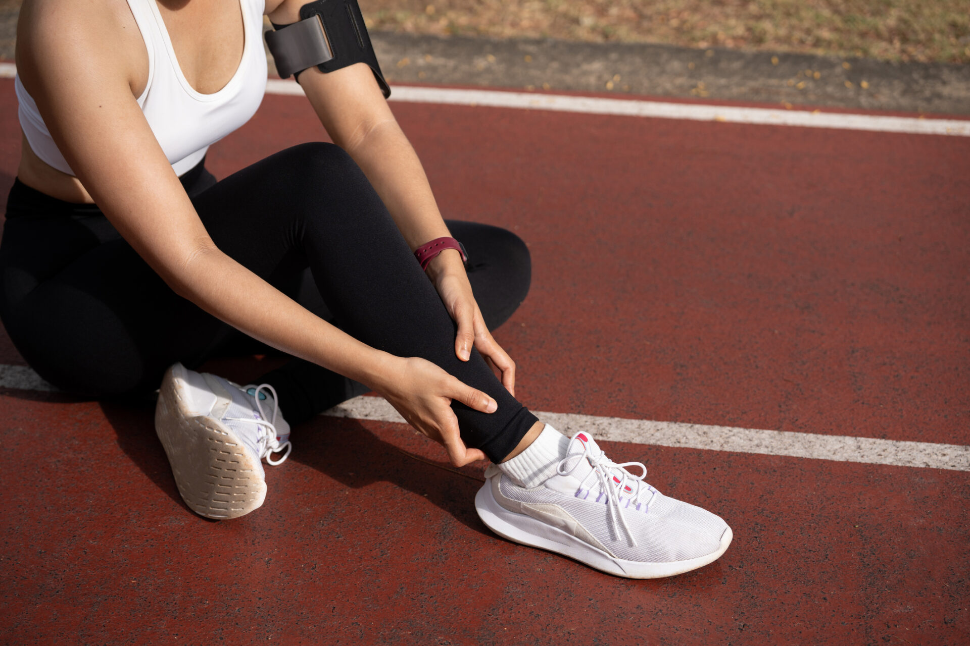 Athlete sits on a red running track, adjusting her black leggings near a white sneaker with a red wristband on her arm.