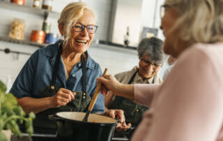 Smiling elderly woman in an apron stirs a pot in a bright kitchen as friends help nearby.