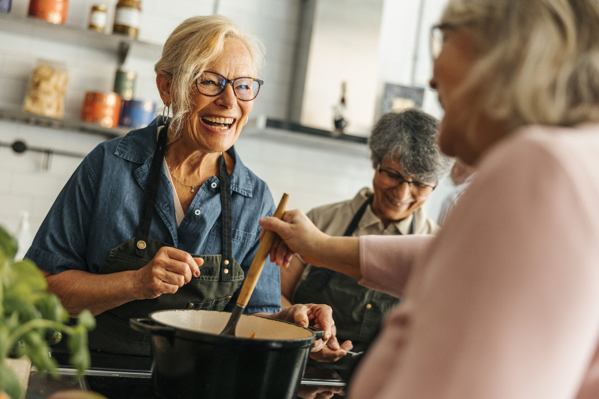 Smiling elderly woman in an apron stirs a pot in a bright kitchen as friends help nearby.