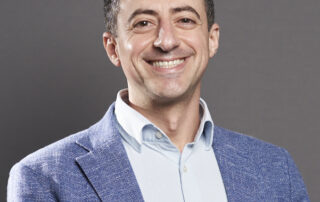 Smiling man in a light blue shirt and blue blazer looking at the camera, professional headshot.