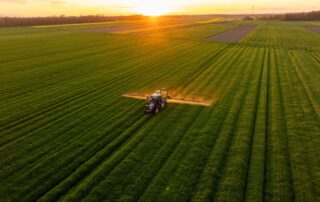 Aerial view of a tractor with a boom sprayer spraying crops across wide, green fields at sunset/sunrise.